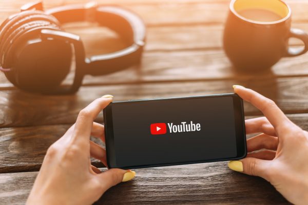 Female hands holding smartphone and using YouTube video service app at wooden table near a cup of coffee and headphones. Point of view in first person.